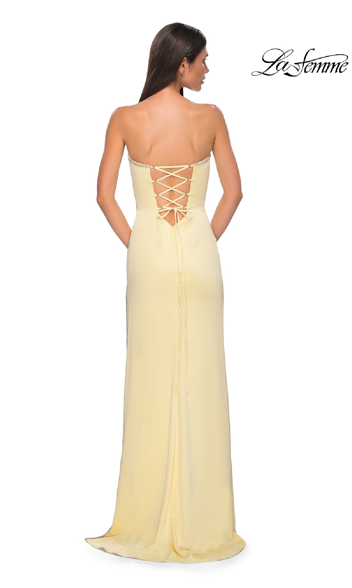 A woman stands facing away, wearing the Classic Strapless Long Prom Dress: La Femme 32799 in light yellow. The dress features a lace-up back detail. The